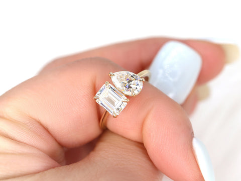 Hand holding two-stone 14kt gold engagement ring with emerald and pear shaped moissanite on light background