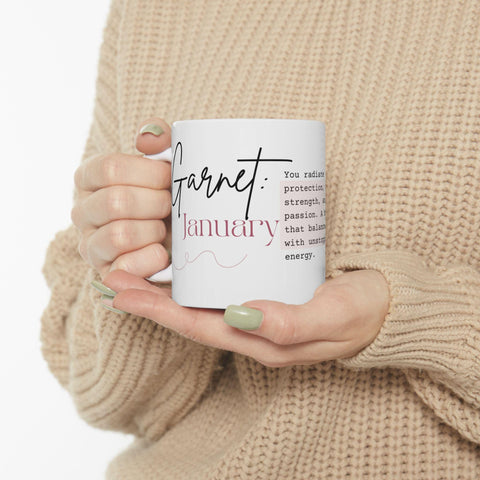 Hands holding white ceramic mug with "Garnet January" text and inspirational message in cozy sweater setting