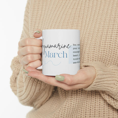 Person holding white ceramic mug with blue script and cursive text on a neutral knit sweater background