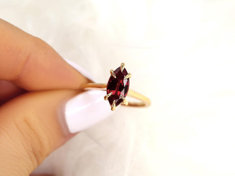 Hand holding a gold ring featuring a marquise-shaped deep red ruby solitaire on a white background