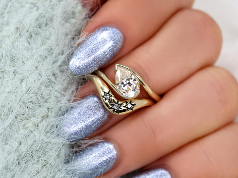 Tilted pear-shaped moissanite bridal ring set with floral engraved gold bands on hand with metallic silver nails