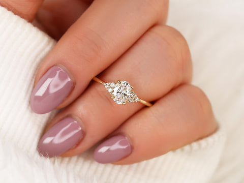 Delicate gold ring with an oval diamond cluster centerpiece worn on a hand with mauve polished nails