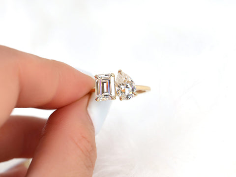 Hand holding a two-stone engagement ring with emerald and pear shaped moissanite on soft white background
