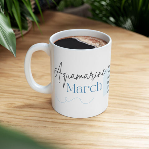 White ceramic mug with "Aquamarine March" text and delicate script filled with coffee on a wooden surface
