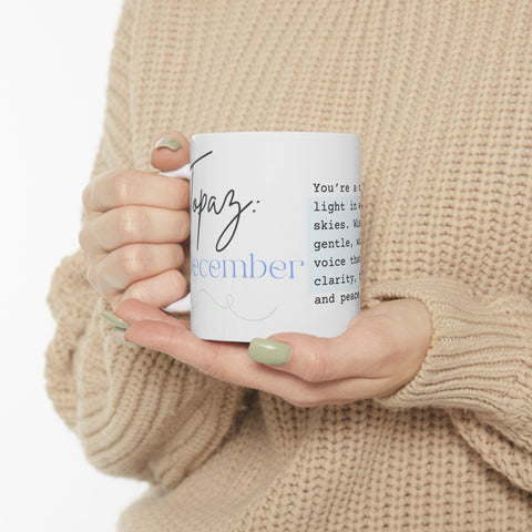 Hands with sage green nails holding a white ceramic mug with blue and black text on a beige sweater background