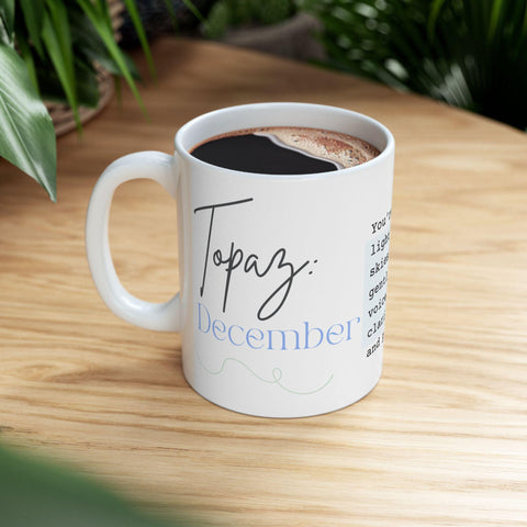 White ceramic mug with "Topaz December" text filled with coffee on wooden surface surrounded by green plants