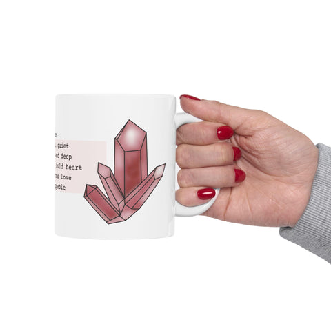 Hand holding white ceramic mug with red crystal illustration and partial inspirational text on a plain background