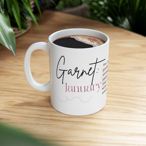 White ceramic mug with "Garnet January" text and decorative script on a wooden table with plants nearby