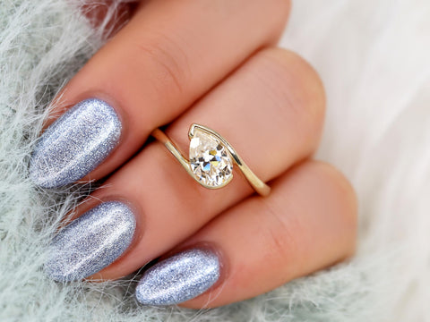 14kt gold ring with a tilted pear-shaped moissanite stone worn on a hand with metallic silver nails